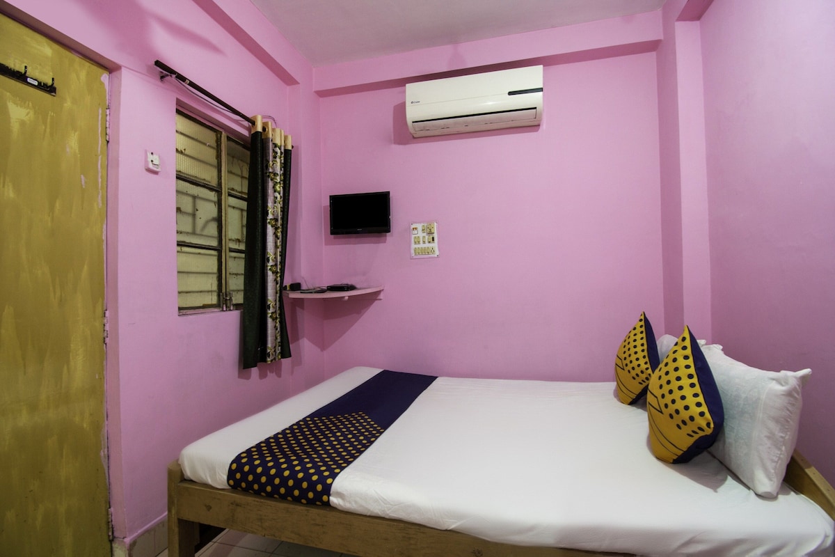 A room with pink walls, a bed with white and patterned bedding, a mounted TV, and an air conditioning unit.