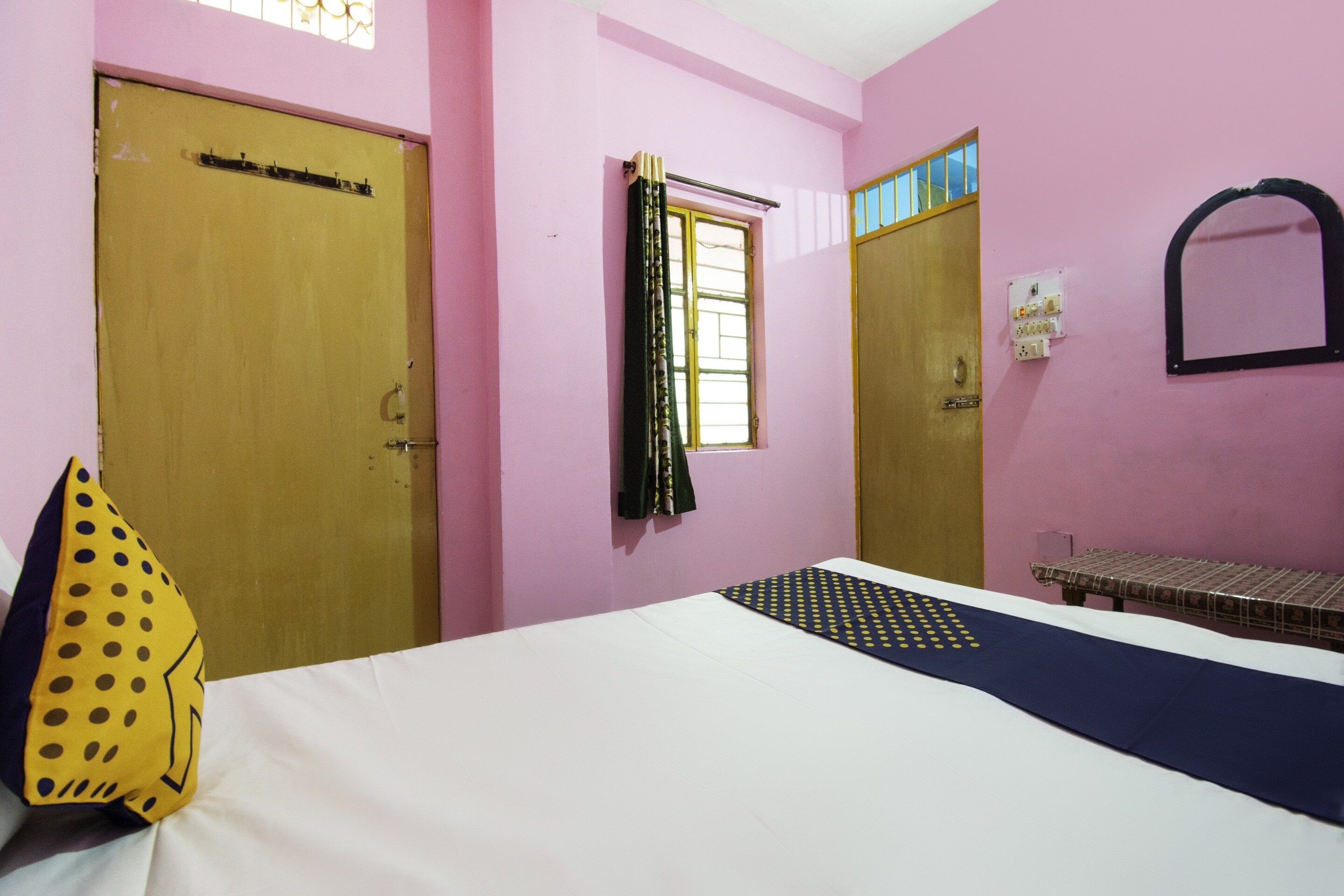 A room with two doors, a window with curtains, a bed with a patterned bedspread, and a small table with a patterned tablecloth.