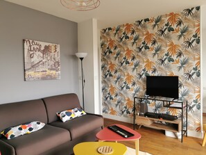 A modern living room with a sofa, a coffee table, a TV on a stand, and a wall with a tropical leaf pattern. - New Listing (REIMS)