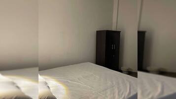 A single bed with a white mattress and pillows, a dark-colored wardrobe, and a wall-mounted light fixture.