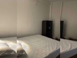 Un lit simple avec un matelas et des oreillers blancs, une armoire de couleur foncée et un luminaire fixé au mur.