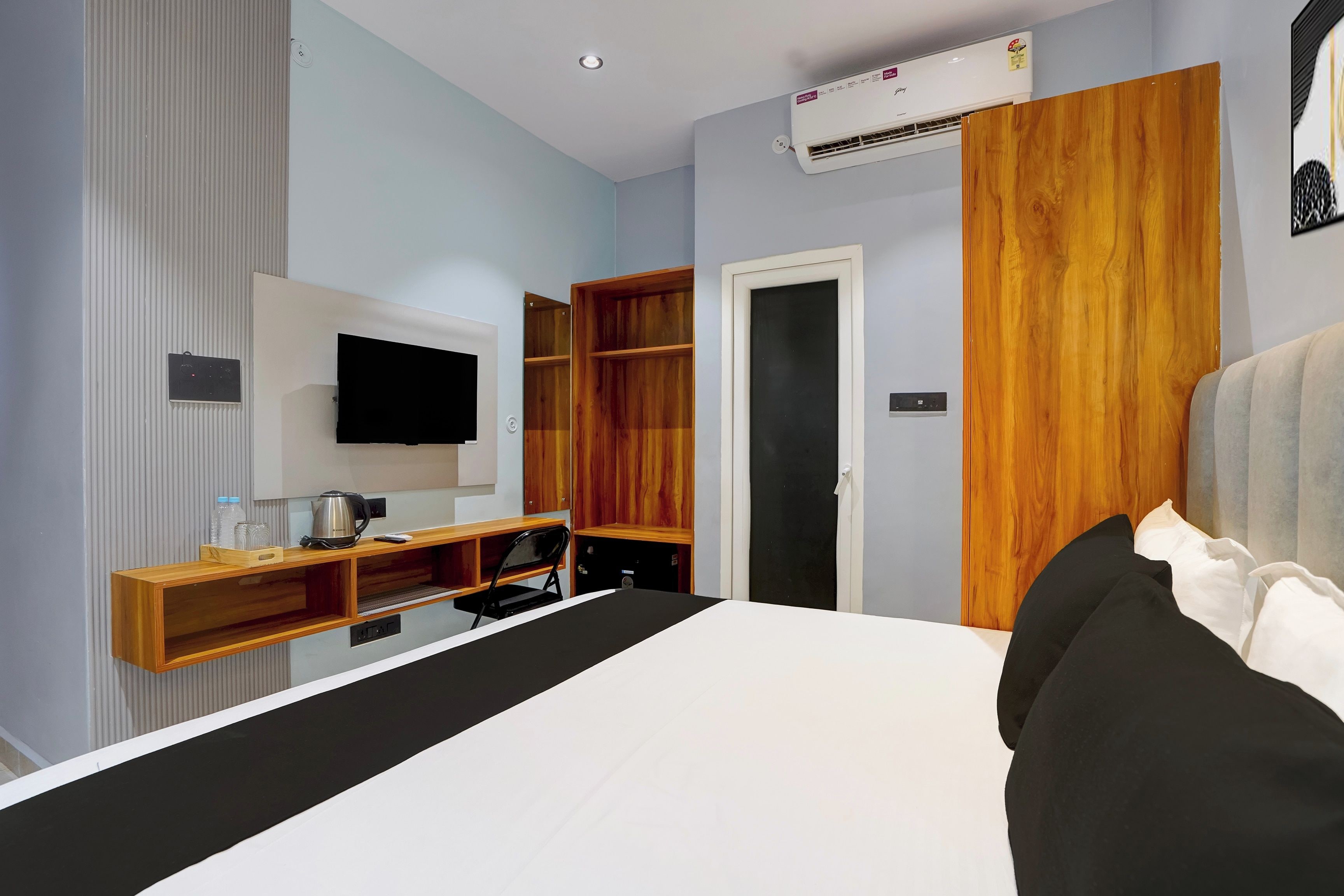 A hotel room with a bed, a television mounted on the wall, a wooden wardrobe, and a built-in desk with a kettle and a bottle.
