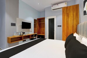 Et hotelværelse med en seng, et fjernsyn ophængt på væggen, en træskab og et indbygget skrivebord med elkedel og en flaske.