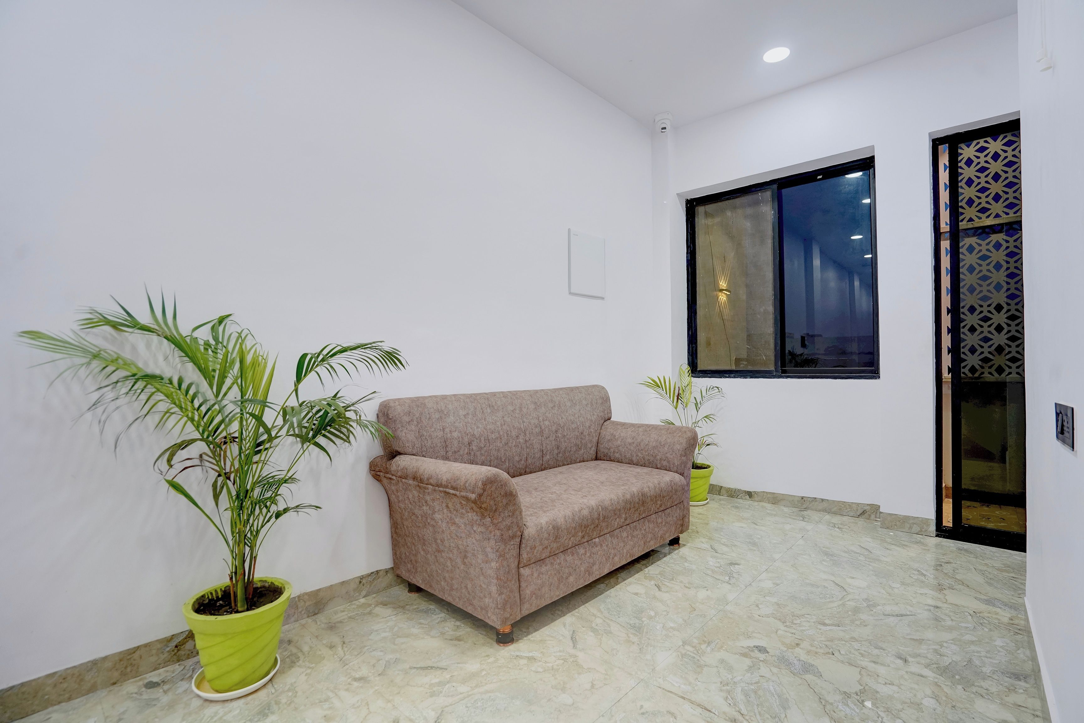 A room with a beige sofa, two potted plants, a window with a geometric pattern, and a marble floor.