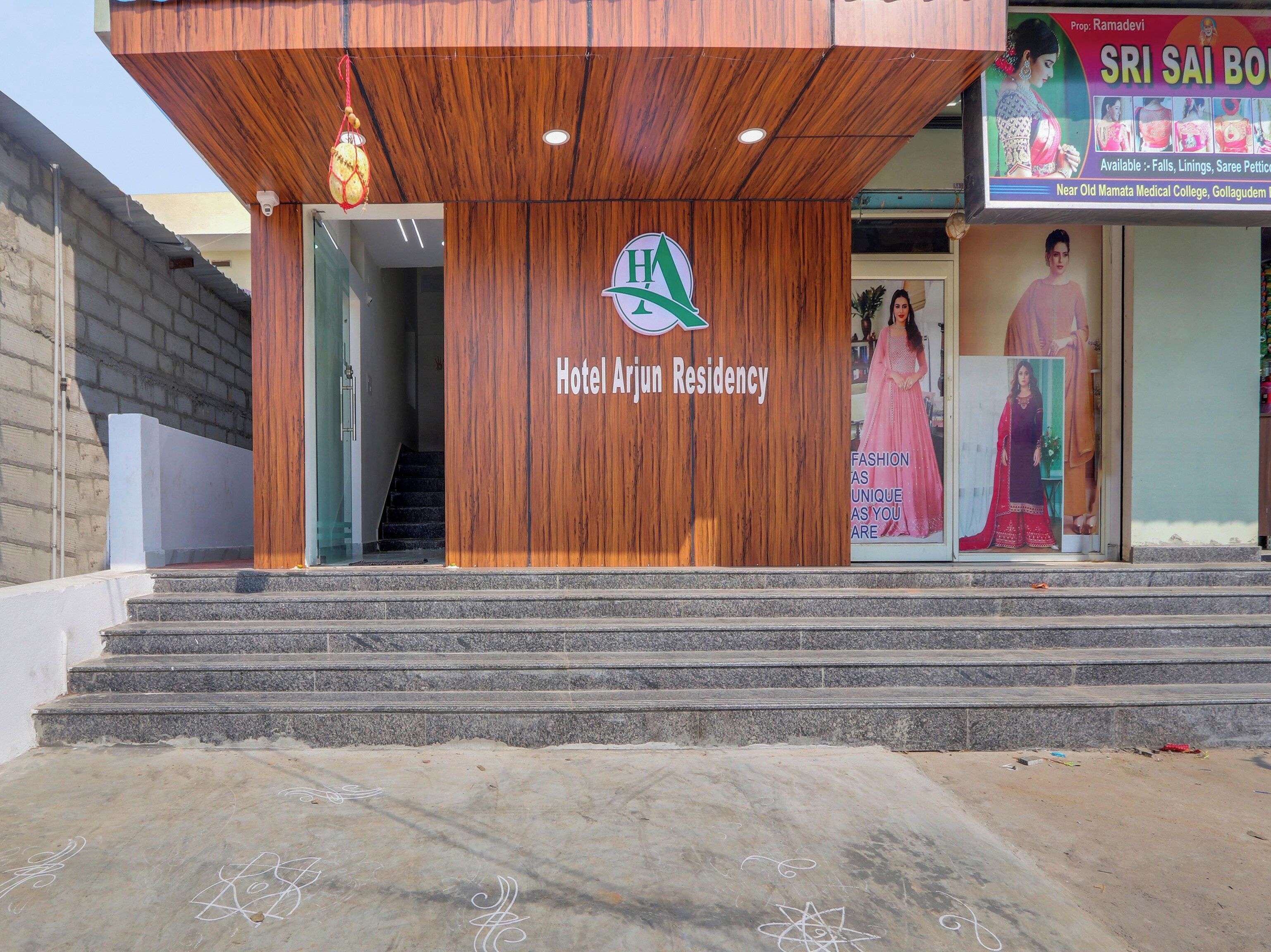 L'ingresso dell'Hotel Arjun Residency, con facciata in legno, una scala che conduce all'entrata e un cartello pubblicitario che offre servizi locali.