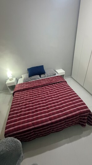 Un dormitorio con cama, mesitas de noche blancas, colcha a cuadros rojo y blanco, y almohada azul.