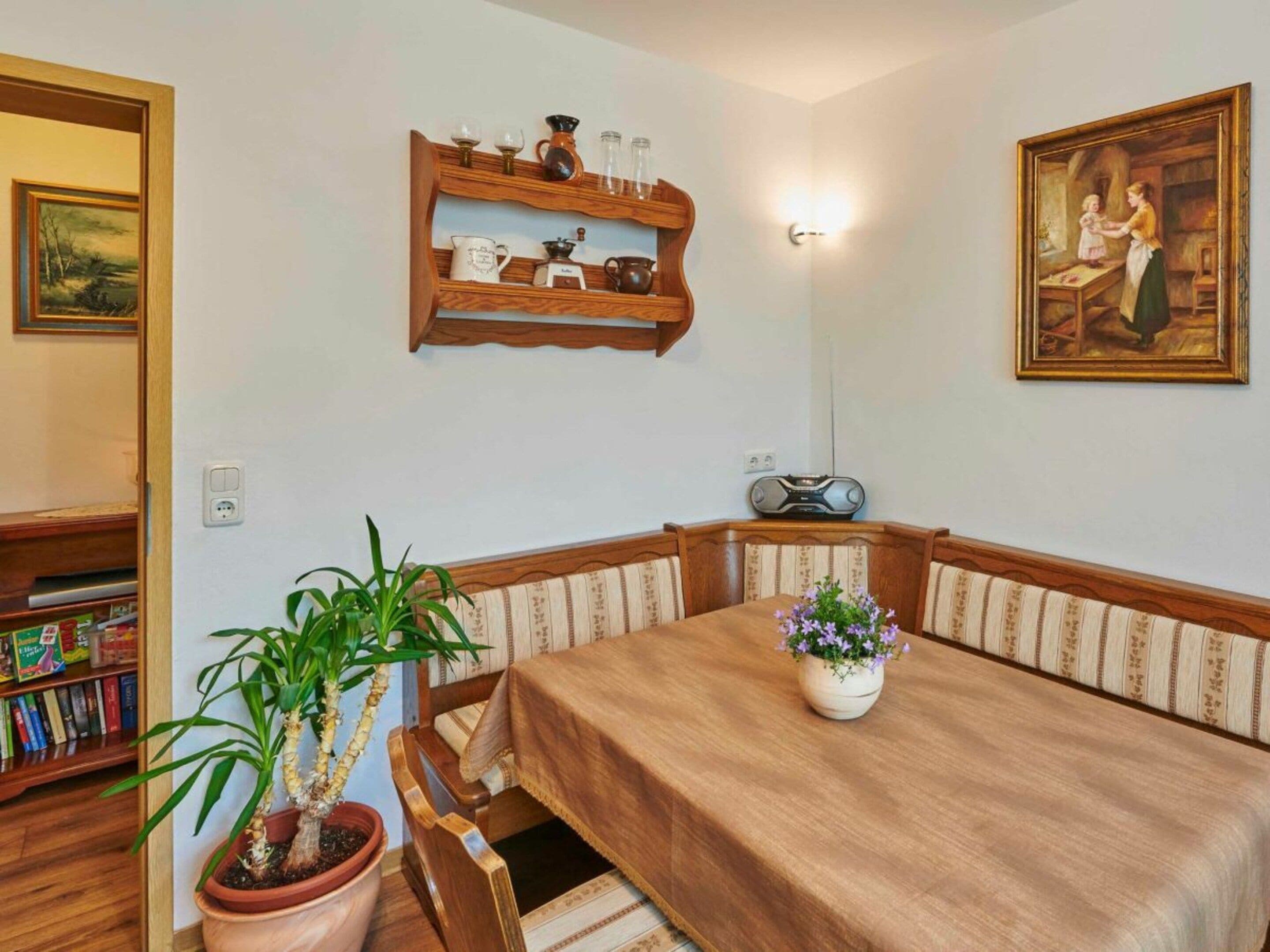 Un comedor con una mesa, sillas, una planta en maceta, un estante con utensilios de cocina y un cuadro en la pared.
