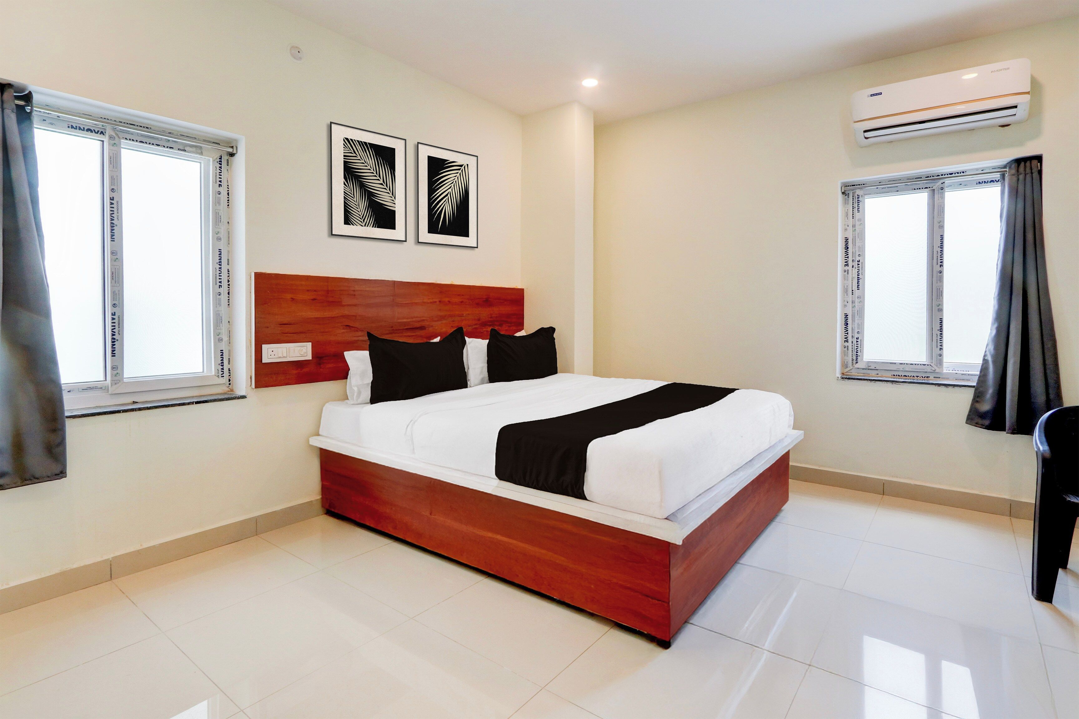 A hotel room with a wooden bed, white bedding, two black pillows, a white bedside table, and a nightstand. There are two windows with curtains, a wall-mounted air conditioner, and two framed leaf artworks on the wall.