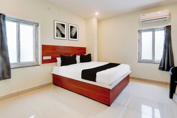 A hotel room with a wooden bed, white bedding, two black pillows, a white bedside table, and a nightstand. There are two windows with curtains, a wall-mounted air conditioner, and two framed leaf artworks on the wall.