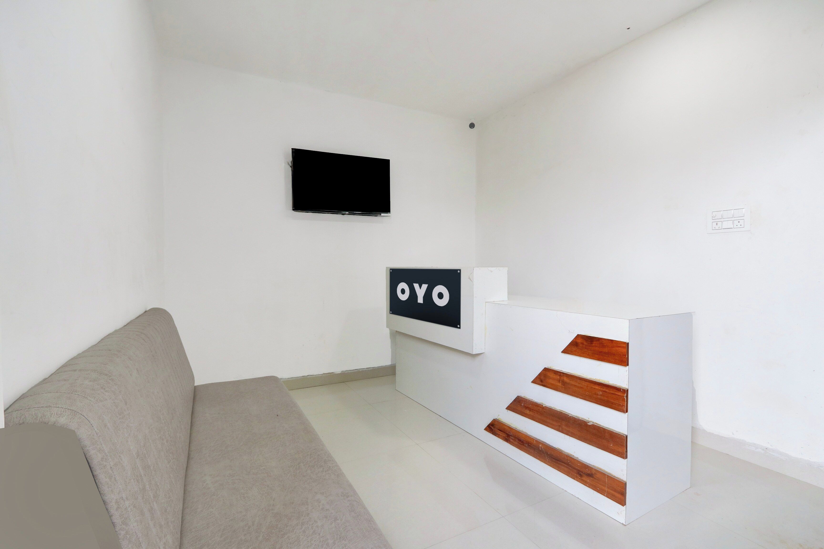 A minimalist room with a grey sofa, a flat-screen TV mounted on the wall, and a reception desk with a sign that reads 'OYO'.