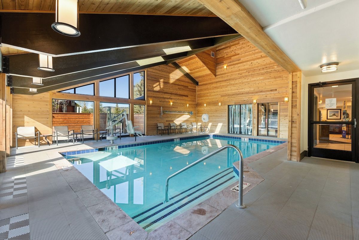 An indoor swimming pool with a wooden interior, a seating area with chairs, and a view through large windows.