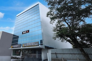 Edificio moderno de hotel con fachada de vidrio, señalización con 'OYO Hotel Grand Delight' y 'Luxury Rooms', y un árbol en primer plano.