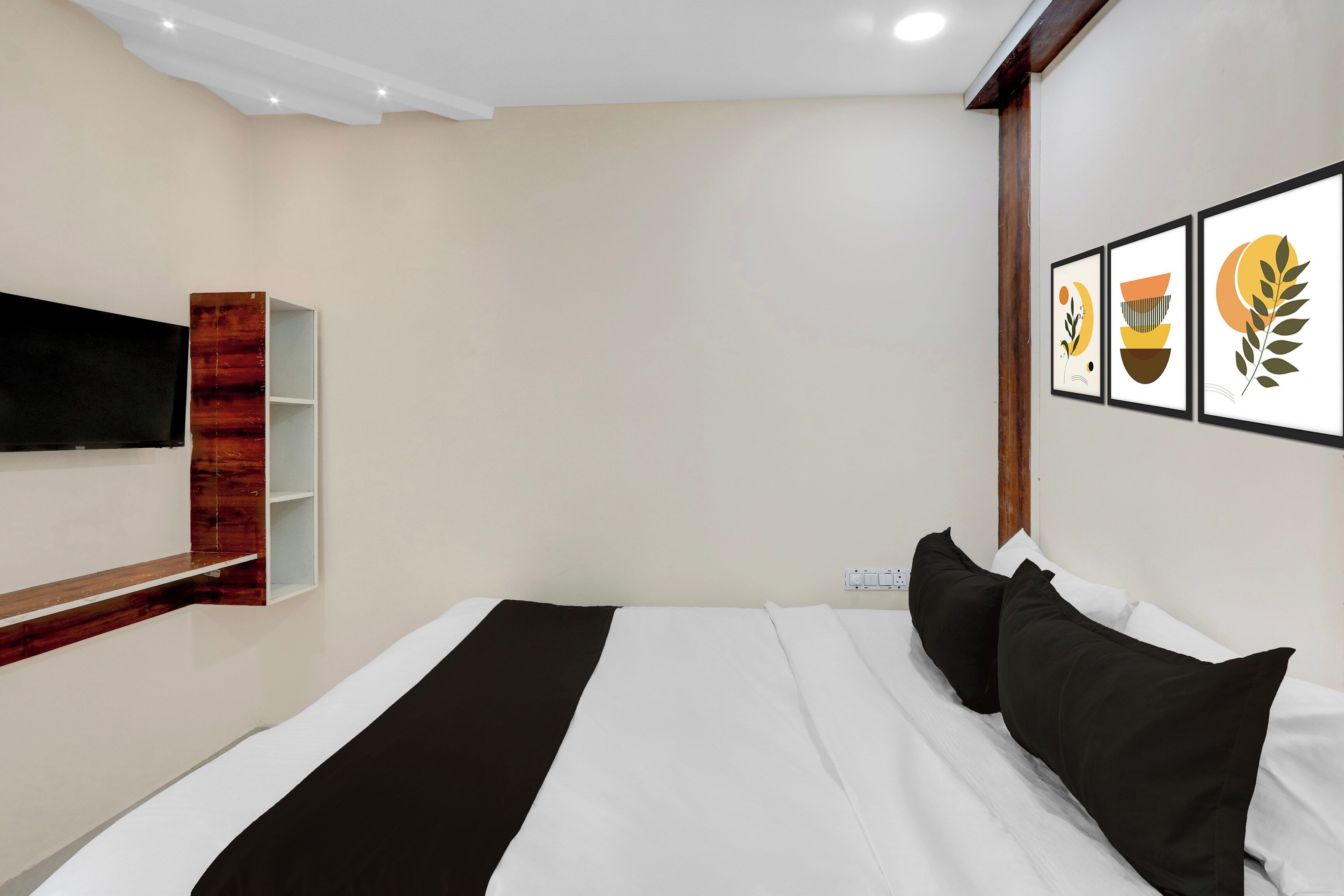 A hotel room with a bed, a mounted TV, a wooden shelf, and three framed artworks on the wall.