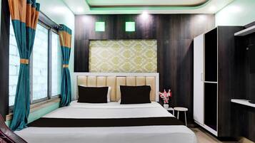 Una habitación de hotel con cama, cortinas, cabecero, una mesita y un armario.