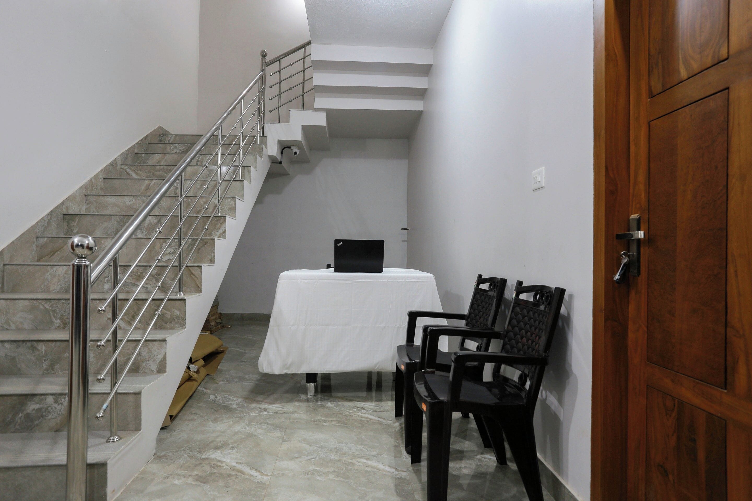 Un escalier avec une rampe en métal, une table blanche avec un ordinateur portable, deux chaises noires et une porte en bois.