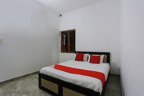Un letto singolo con struttura in legno e materasso bianco, corredato da cuscini rossi e bianchi, in una stanza dalle pareti bianche e con una piccola finestra.
