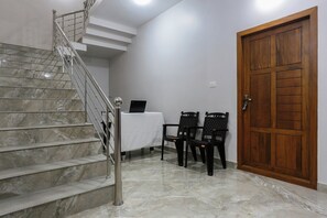 A staircase with a metal railing, a wooden door, and two black chairs with a table in a room with white walls and marble flooring.