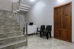 Una escalera con barandilla de metal, una puerta de madera y dos sillas negras con una mesa en una habitación de paredes blancas y suelo de mármol.