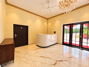 Un hall d’hôtel avec un comptoir d’accueil, une porte sombre, un ventilateur de plafond et une grande fenêtre donnant sur un paysage verdoyant.