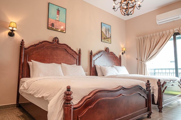 A double bed with a wooden frame and white bedding in a room with beige walls, two paintings, a chandelier, and an air conditioning unit. - The Farmhouse (Ipoh)