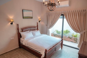 A bedroom with a wooden bed, two wall-mounted lamps, a ceiling light, a painting, and a view of the outdoors through a window with curtains. - The Farmhouse (Ipoh)