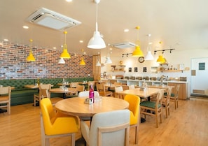 A restaurant interior with wooden tables, yellow chairs, and a brick wall. - Bloom Hotel - Jayanagar (Bengaluru)
