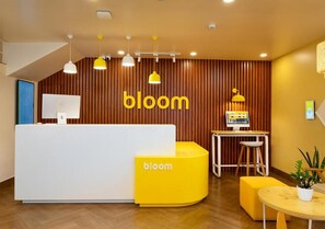 A reception area with a 'bloom' sign, a white reception desk, a yellow seating area, and a small table with a plant. - Bloom Hotel - Jayanagar (Bengaluru)
