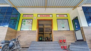 Entrada d'un edifici amb façana groga, un rellotge i indicadors amb la lectura 'SRI SAKTHI', 'BENVINGUT' i 'MAHAL'.