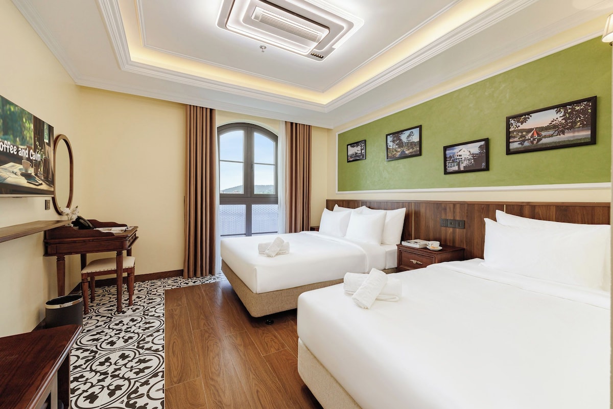 Ein Hotelzimmer mit zwei Betten, einem Schreibtisch, einem Stuhl, einem großen Fenster und einer grünen Wand mit gerahmten Bildern.