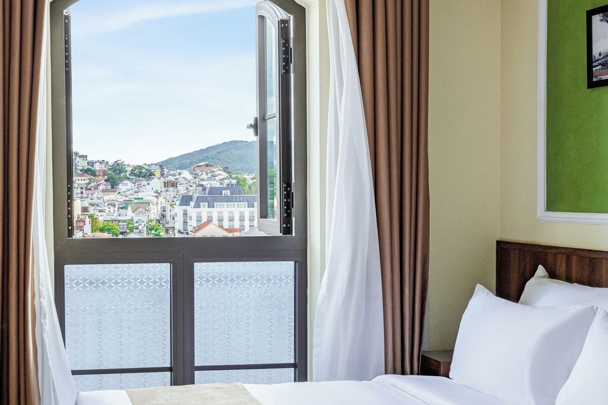 Ein Hotelzimmer mit einem großen Bett, Blick auf eine Stadtlandschaft durch ein Fenster und einer grünen Wand.