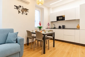 A modern kitchen with white cabinets, a microwave, and a coffee maker. There is a dining table with chairs and a blue sofa in the living area. - New Listing (ROMA)