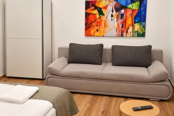 A modern room with a sofa, a bed, a remote control on a side table, and a colorful abstract painting on the wall. - Curtain Call Vienna 1BR near Stadthalle (Vienna)