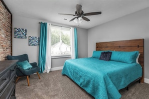 Ein Schlafzimmer mit einem Bett, einem Sessel, einem Schminktisch, einem Deckenventilator und einem Fenster mit Vorhängen.