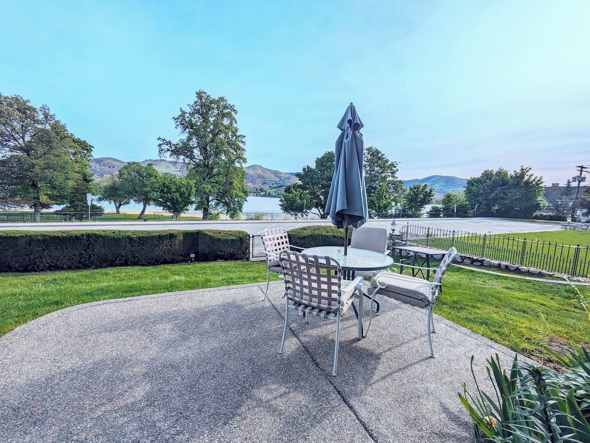 En udendørs terrasse med et rundt bord og stole, en grå parasol og udsigt til en park med træer og fjerne bakker.