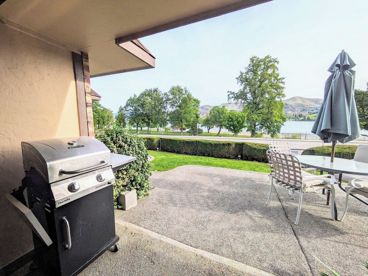 An outdoor patio with a barbecue grill, a table with chairs, and a view of a park and hills.