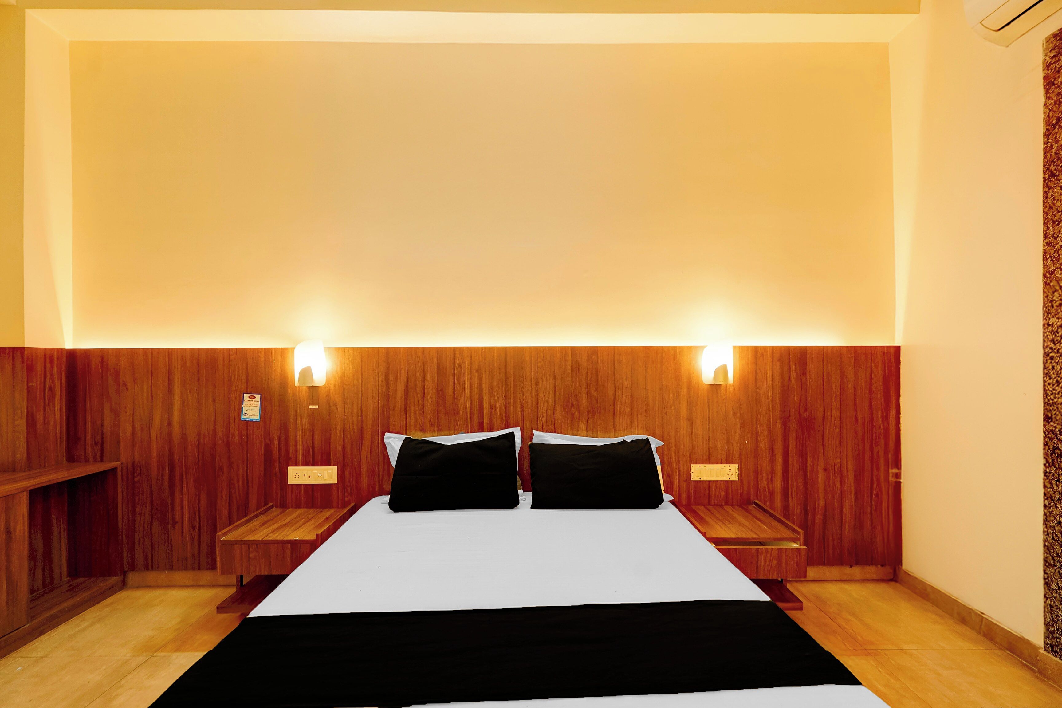Una habitación de hotel con cabecera de madera, dos mesitas de noche, una cama con ropa de cama blanco y negro, e iluminación montada en la pared.