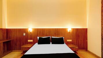 Una habitación de hotel con cabecera de madera, dos mesitas de noche, una cama con ropa de cama blanco y negro, e iluminación montada en la pared.