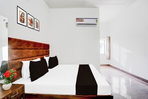 Une chambre d’hôtel avec un lit, une table de chevet, un vase de fleurs, des œuvres d’art encadrées au mur et un appareil de climatisation.
