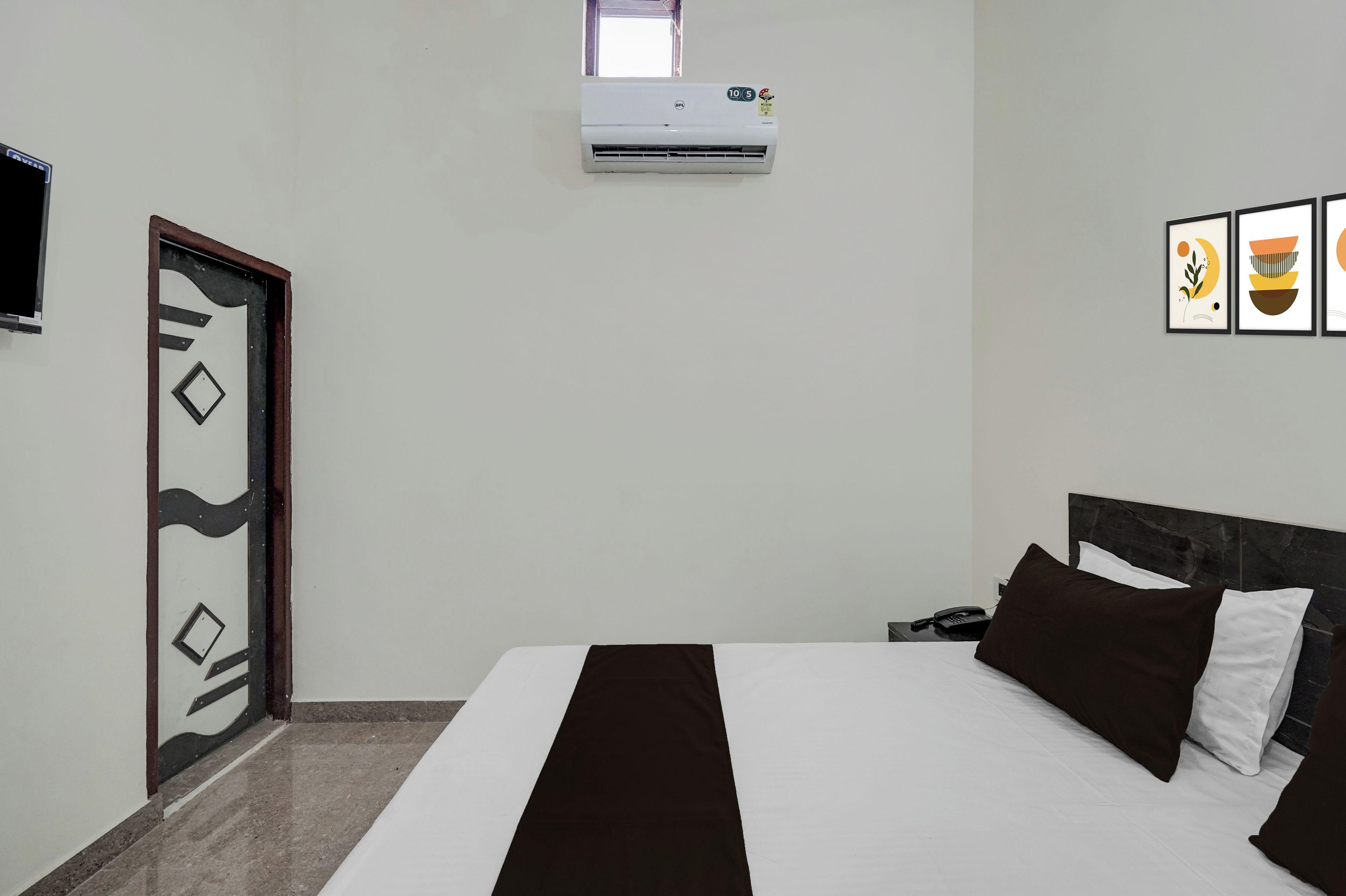A hotel room with a bed, a desk with a phone, an air conditioning unit, a door with a geometric design, and framed artwork on the wall.