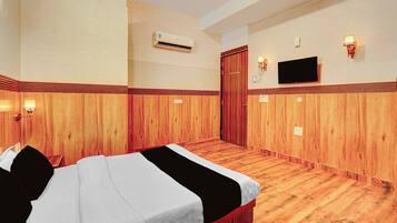 Habitación de hotel con piso de madera, cama con ropa blanca, televisor montado en la pared y equipo de aire acondicionado.
