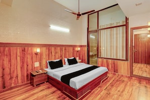 A room with a wooden floor, a bed with a white bedspread and black pillows, a nightstand with a lamp, and a large window with curtains. - HOTEL O SAFFRON (Meerut)