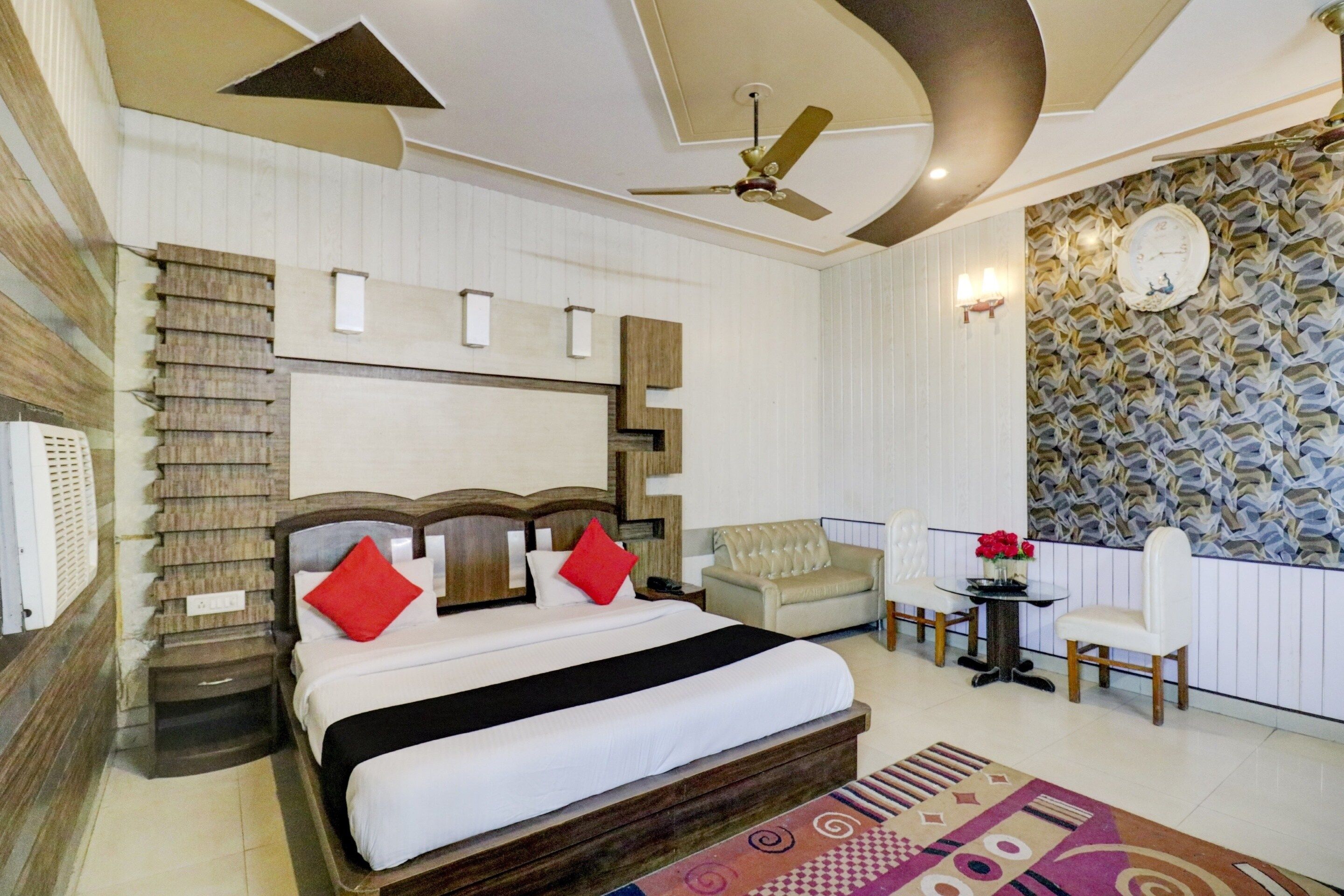 Habitación de hotel con una cama grande, un ventilador de techo, un papel tapiz estampado y una zona de estar con mesa y sillas.