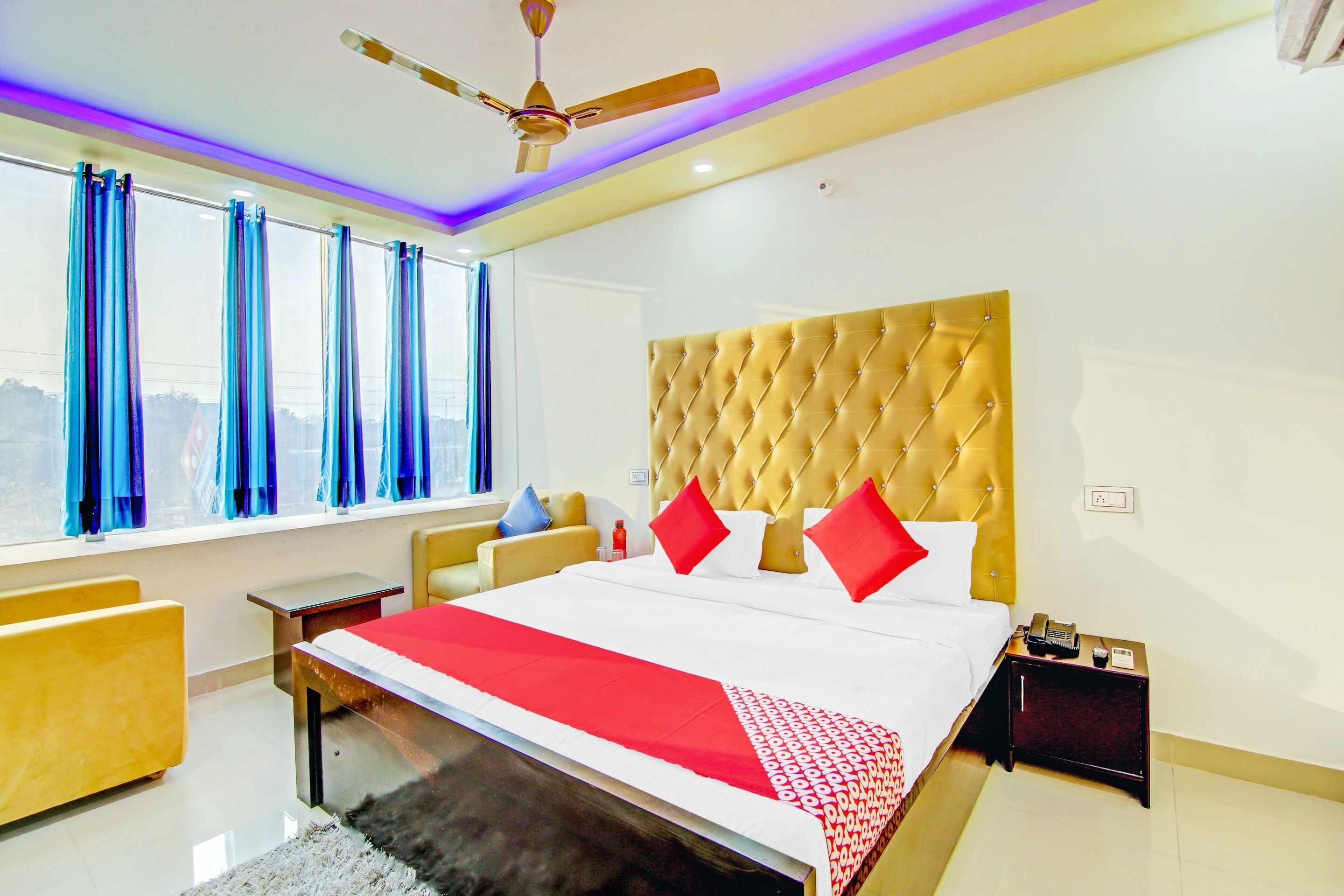 Quarto de hotel com cama grande, ventoinha no teto, mesa de cabeceira e janela com cortinas.