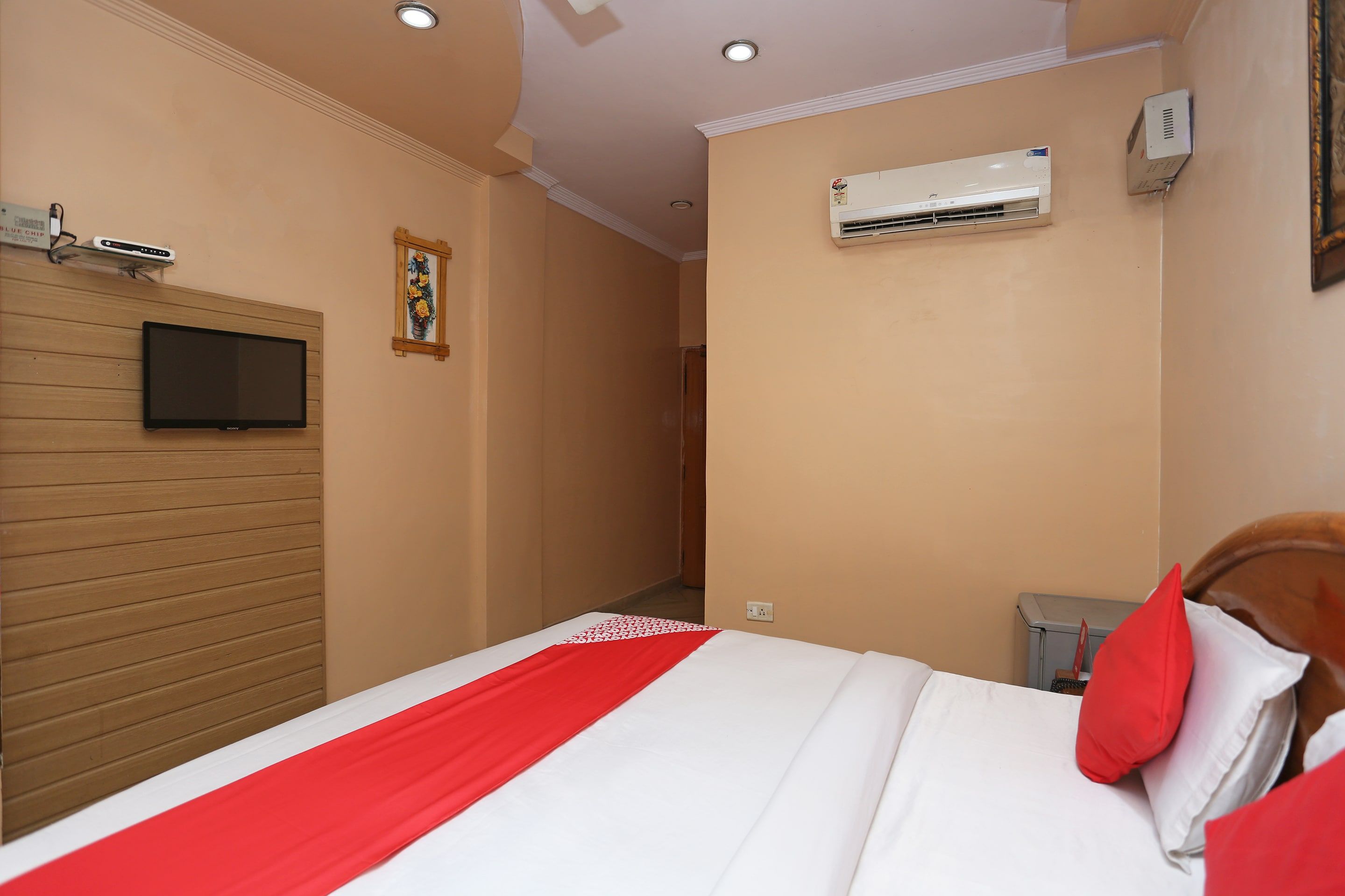 Habitación de hotel con cama, televisor montado en un panel de madera, equipo de aire acondicionado y un enchufe en la pared.