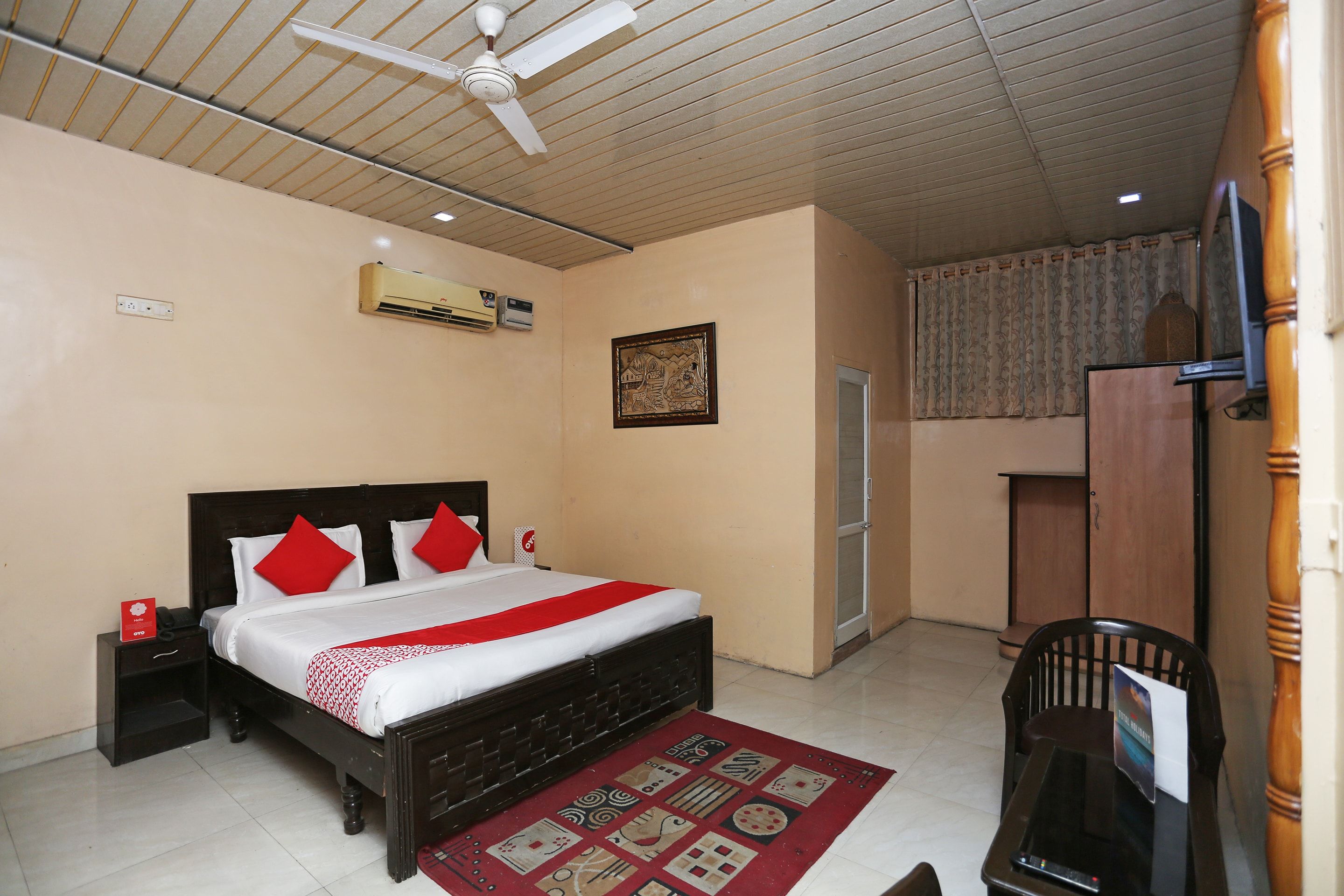 A hotel room with a bed, a desk, a chair, a ceiling fan, an air conditioning unit, and a window with curtains.