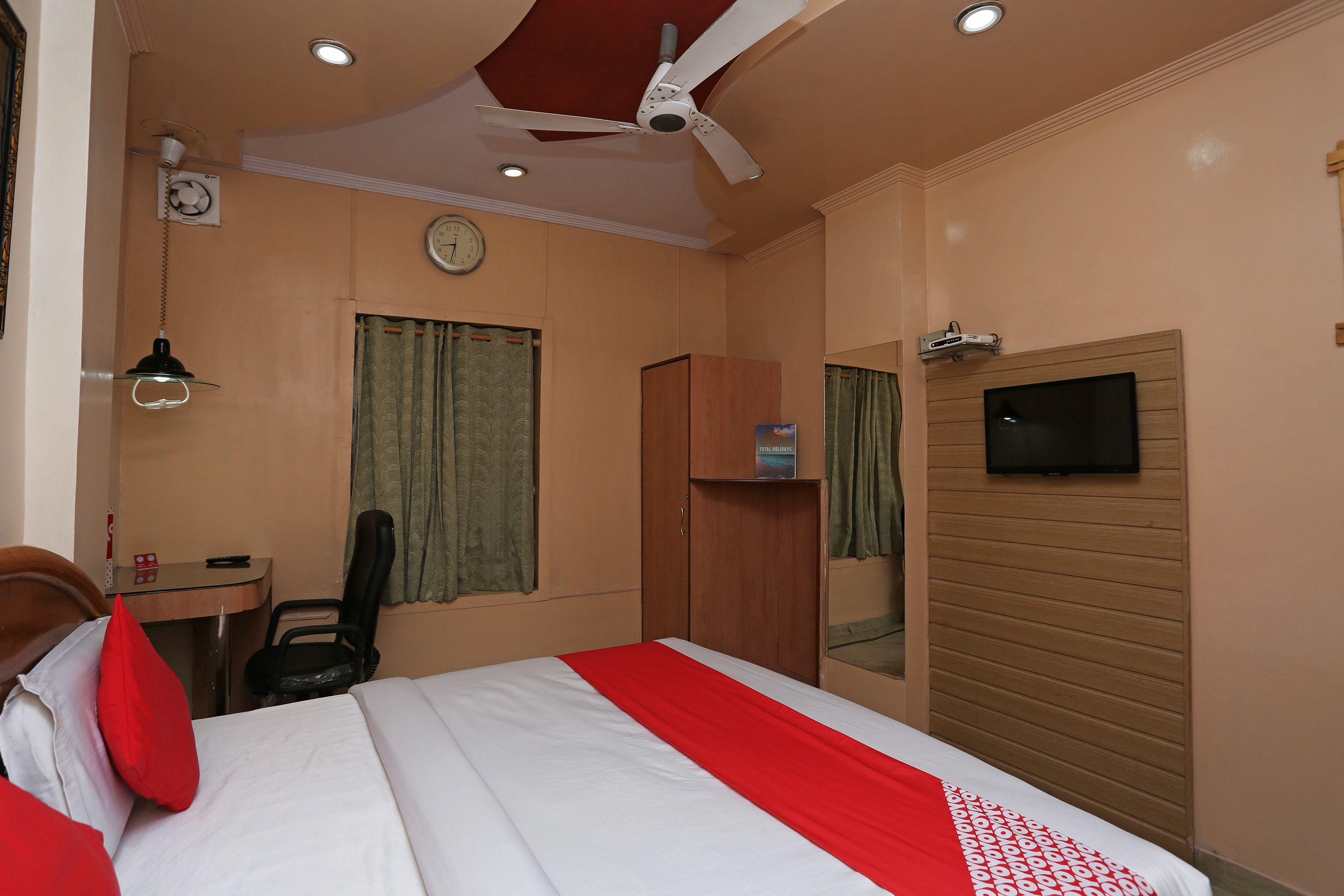 A hotel room with a bed, a desk, a chair, a wardrobe, a television, and a ceiling fan.