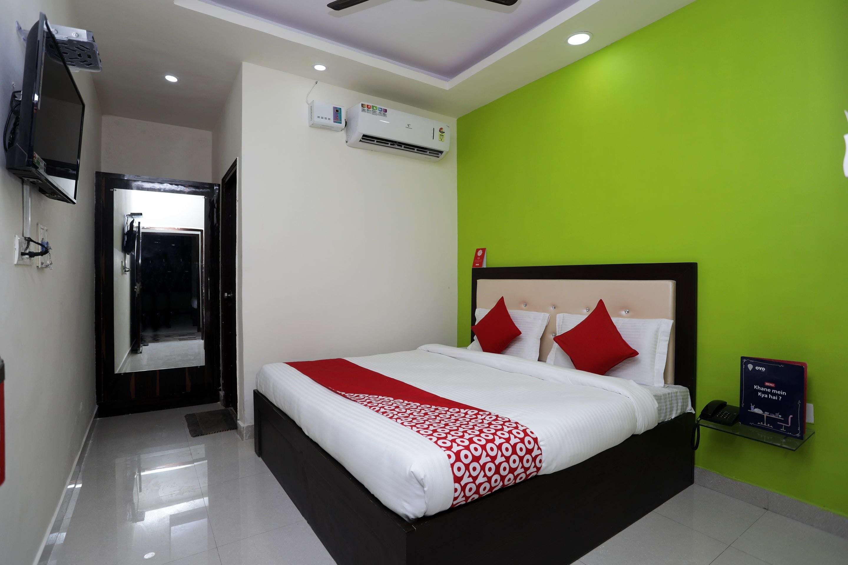 A hotel room with a bed, a TV, an air conditioning unit, and a green wall.