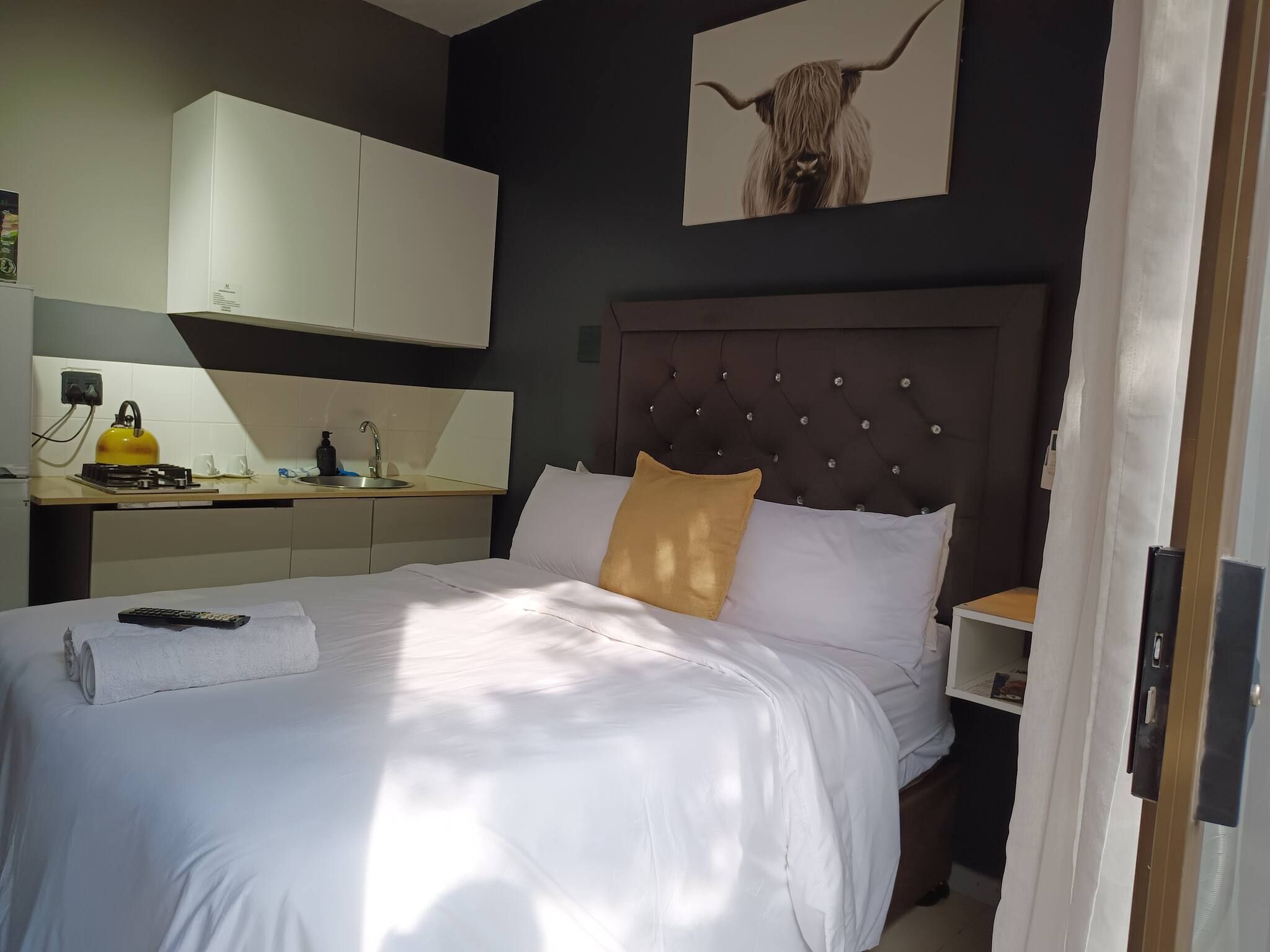 A hotel room with a bed, a bedside table, a nightstand, a wall-mounted artwork, and a kitchen area with a kettle and a sink.