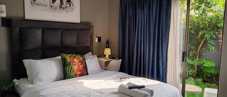 A hotel room with a bed, a bedside table with a lamp, a wall-mounted air conditioner, and a large window with curtains.