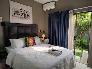 A hotel room with a bed, a bedside table with a lamp, a wall-mounted air conditioner, and a large window with curtains. - Durban Cozy Stay - Free Wifi & Parking (Durban)